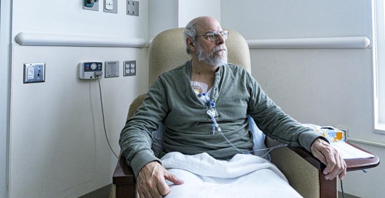 A recovering senior adult man colorectal cancer patient is sitting resting comfortably in a hospital cancer ward easy chair while chemotherapy IV drip medicine is administered by an array of medical equipment through a subcutaneous intravenous chemo access port temporarily embedded into his upper chest. 