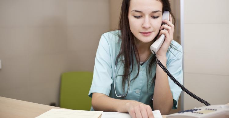 Young female nurse sitting at a desk speaking over the phone while looking at papers