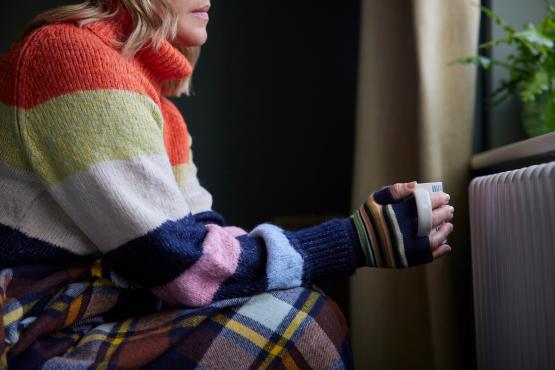 Woman In Gloves With Hot Drink Trying To Keep Warm By Radiator During Cost Of Living Energy Crisis