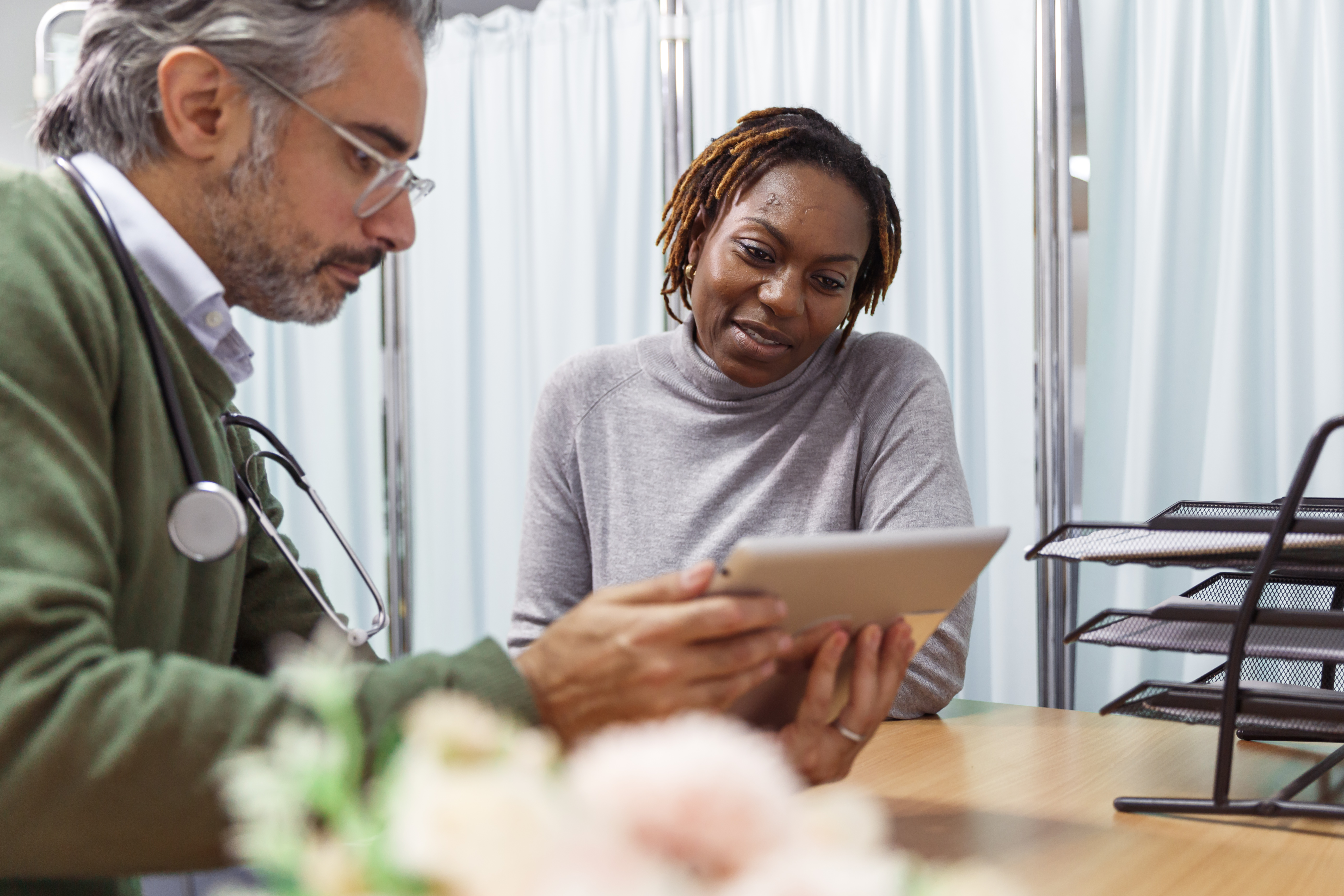 A black female patient reviews medical test results with her male doctor who is of Middle Eastern descent during a consultation. They are seated next to each other in a medical office and the doctor is holding a tablet computer.