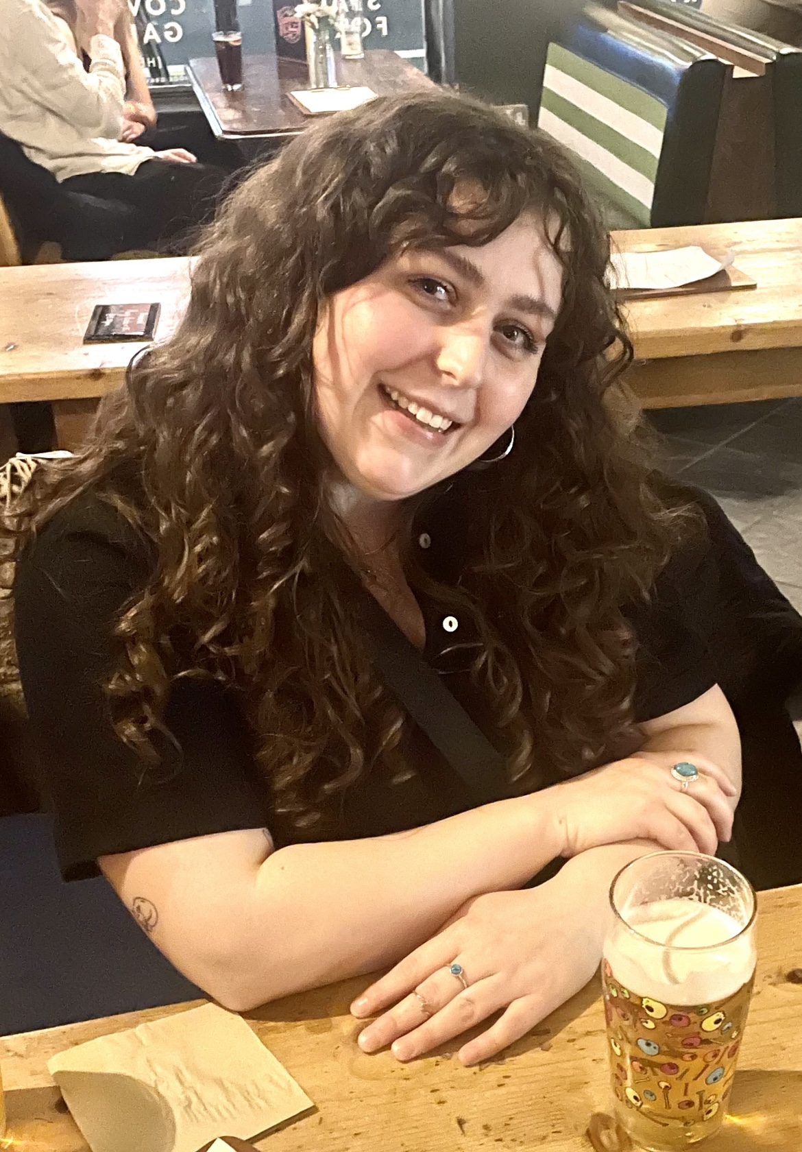 A white woman with long wavy dark hair sits in a pub with a pint of beer in front of her.