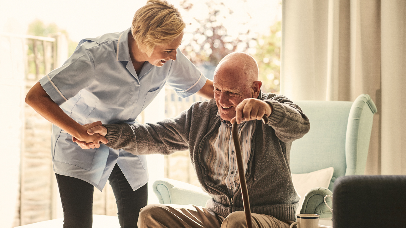 A female nurse is helping an elderly man out of a chair