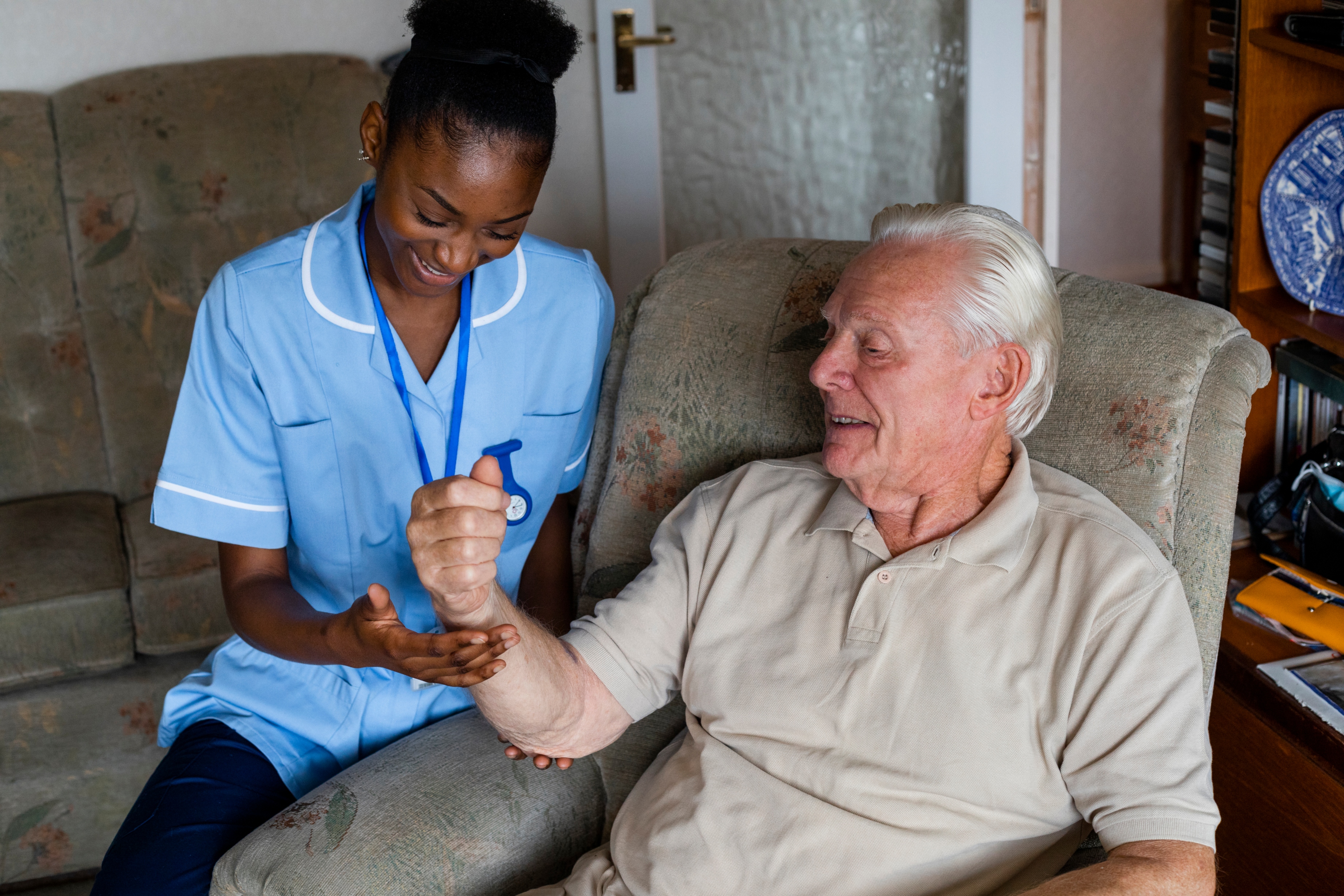 Nurse examining/treating a senior patient with arthritis who is sitting in a chair in his living room in the North East of England. The nurse is looking over his hand.