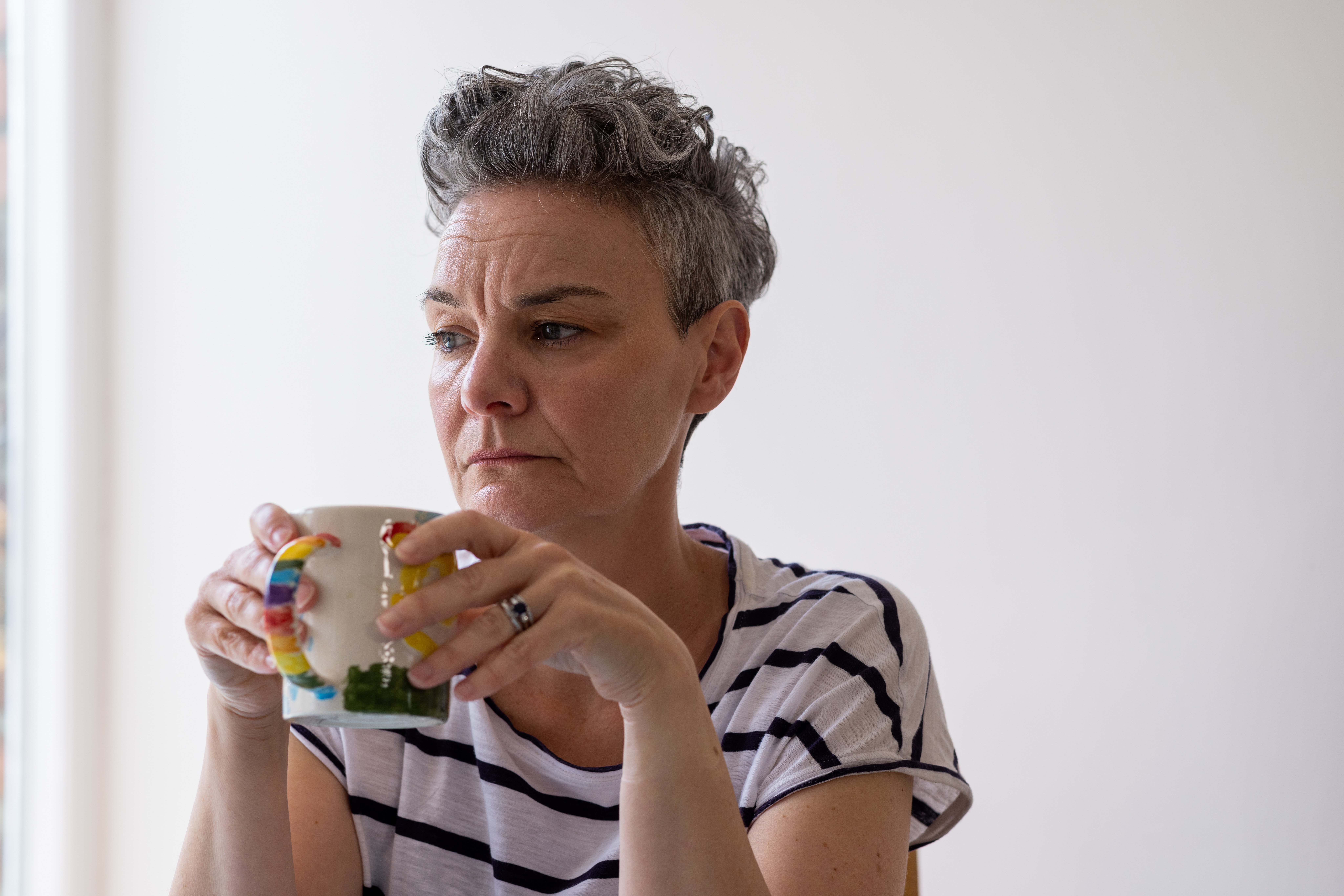 Close-up of a mature female sitting at a table in a kitchen having a cup of tea with a negative emotion on her face. She is taking time to herself overthinking personal issues.