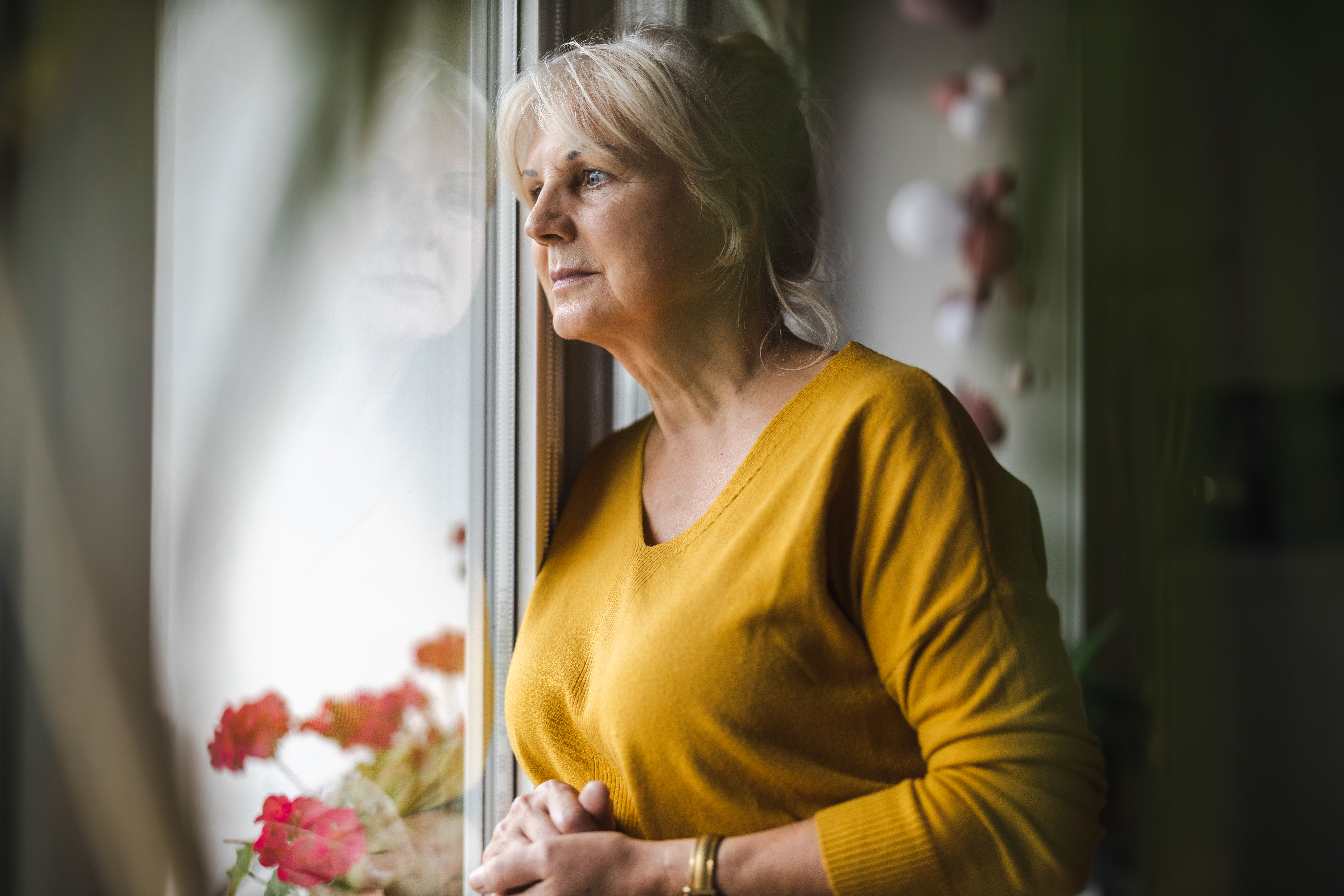 An older woman wearing a yellow jumper her blonde hair in a ponytail stands looking worriedly out of a window, clasping her hands together