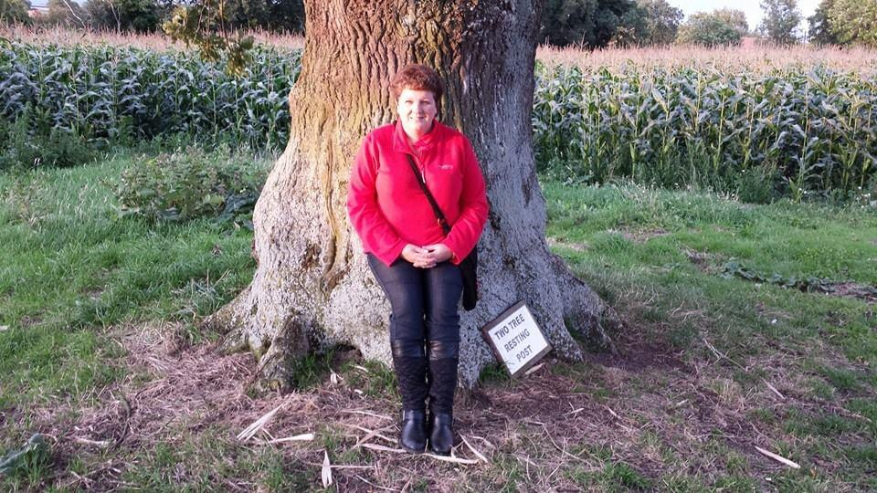 Sue is a white woman wearing a red top and leaning against a tree.