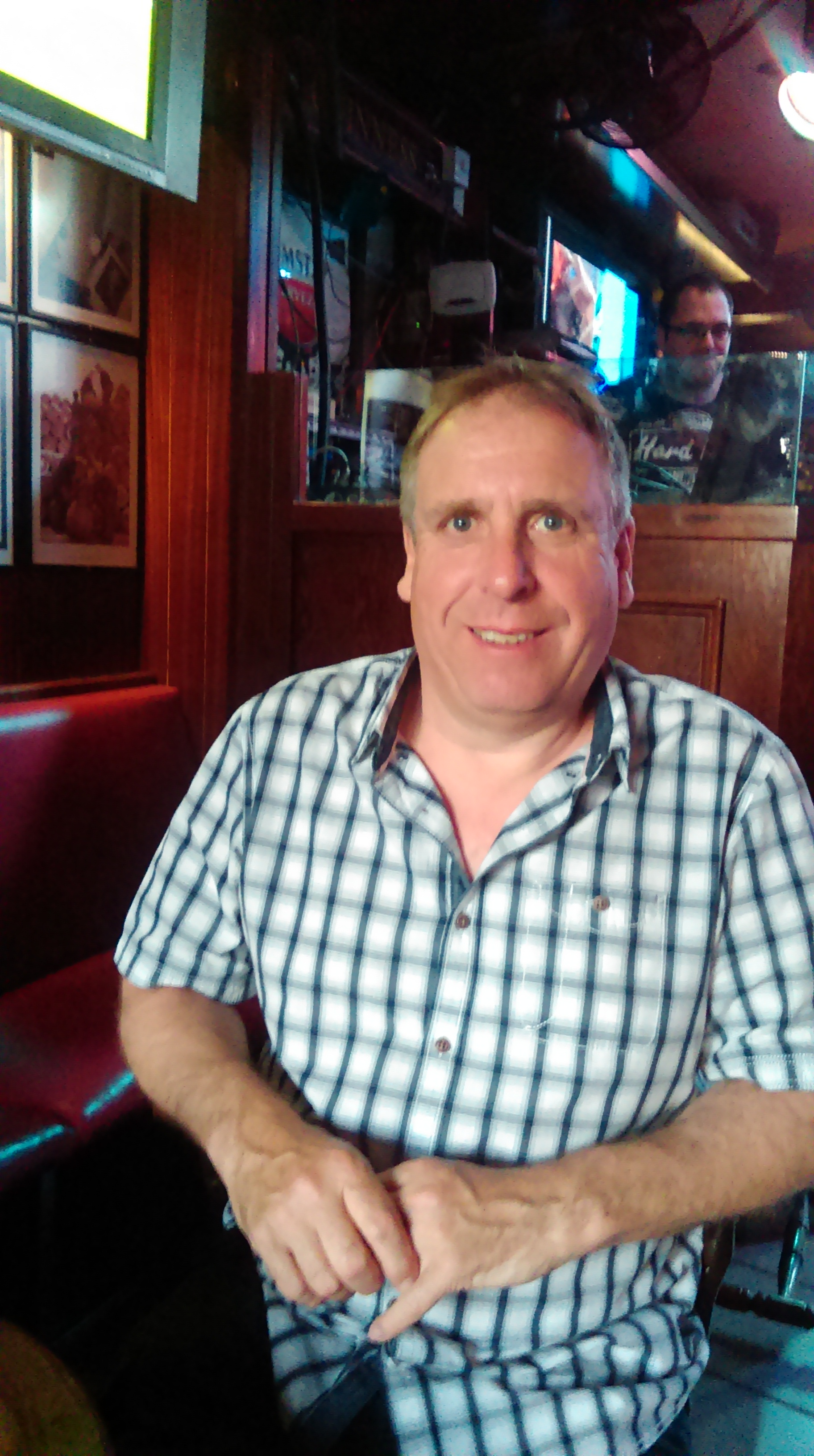 Alan is a white man with grey hair. He is sitting in a wood panelled pub in a green and white checkered shirt with the top button open.