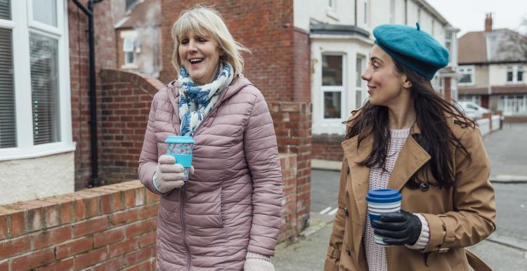 Two women drinking hot drinks in reusable cups walking down a street together, talking and catching up.