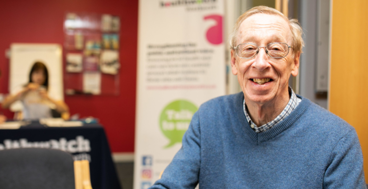 An elderly white man is sitting in front of a Healthwatch information sign. He is wearing glasses and a blue jumper and is smiling.