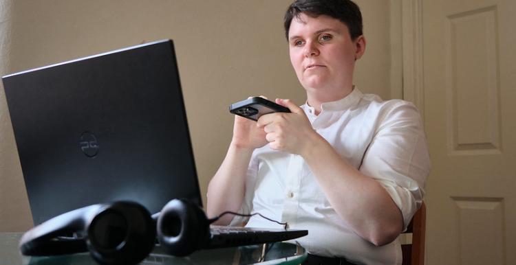 A visually impaired person is sitting at a computer. They are using a screen reader on their mobile phone. They are wearing a white shirt and black trousers.