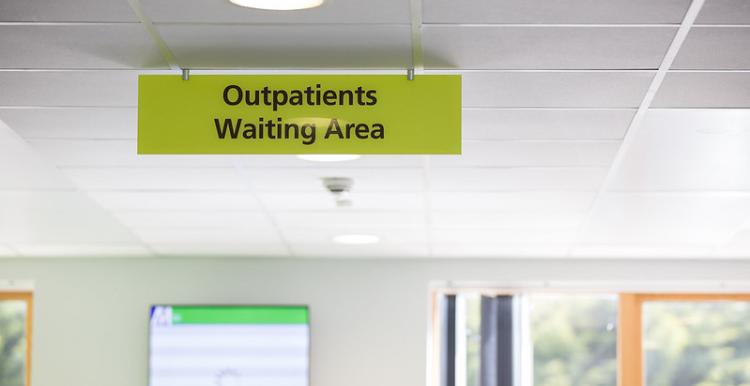 A clinic sign on a green background with black text says 'outpatients waiting area'. There is a window and information board in the background.