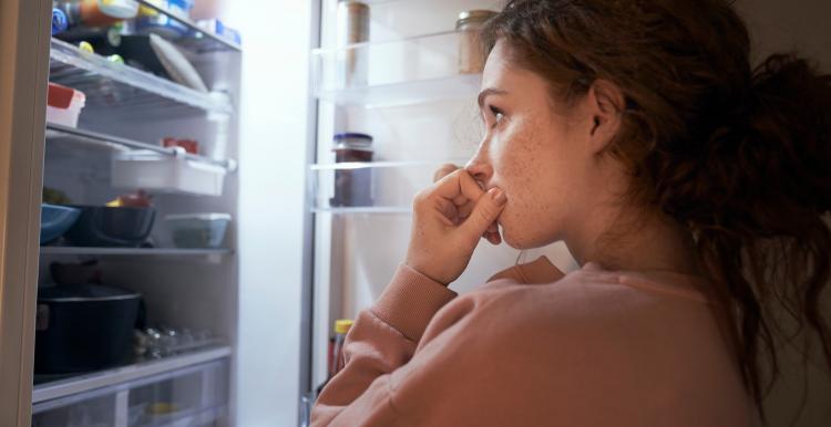 A young woman with a worried expression looks into a fridge filled with food