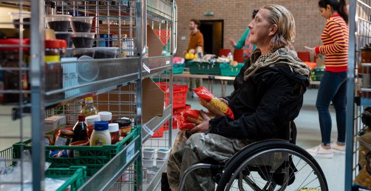 Side view of a volunteer in a wheelchair organising food donations into storage at a food bank in the North East of England. Behind him are his co workers who are working out of focus