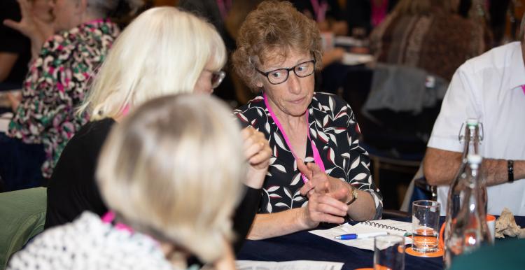 Jane Laughton is a middle-aged white woman. She is wearing glasses, a black and white shirt and has short, light-brown, curly hair. She is talk to other memebers of Healthwatch at a conference.