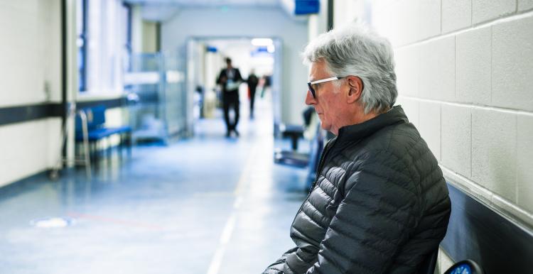 Portrait of a senior man in his 70s sitting and waiting in a hospital corridor. Focus on the man with a worried and anxious expression on his face, while the background of the hospital is defocused.