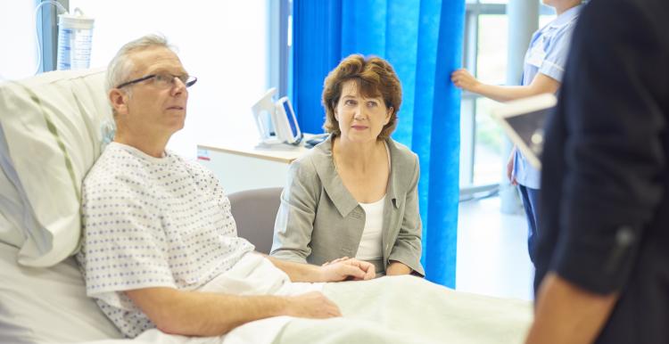A mature man sits in his hospital bed waiting to listen to a doctor explain his diagnosis whilst being consoled by his wife . In the background a young nurse adjusts his curtain screen so that he can hear the news in private .