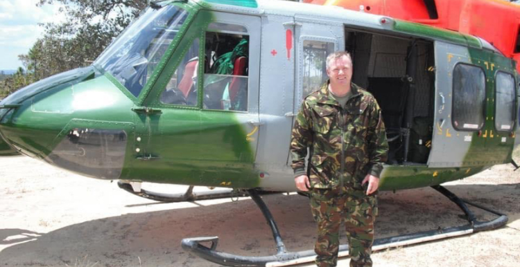 Shaun is wearing a camouflage uniform and standing in front of a helicopter.