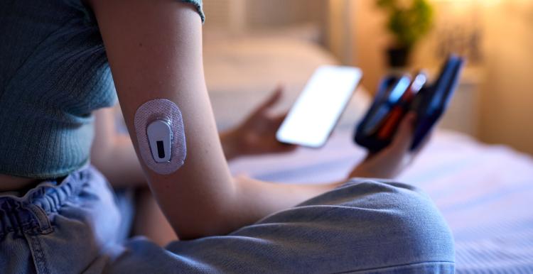 A person is sitting on a bed. They are looking at diabetes monitoring equipment and a mobile phone. They have a monitoring device on their right arm.
