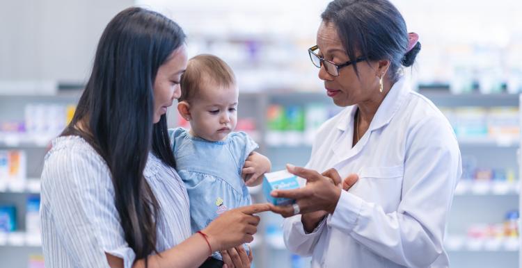 A young mother of Asian descent is picking up a prescription. The woman is holding her baby girl while at the store. She is speaking to a female pharmacist of African descent about the medicine. The medical professional is holding the box of medicine.