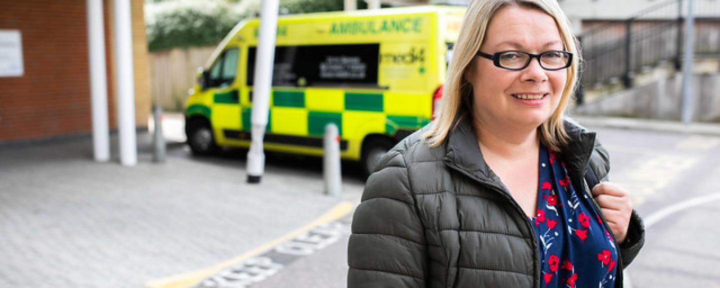 A blonde woman in a coat, standing in front of an ambulance 