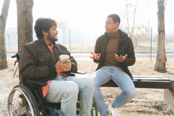 Two men sit outside, one is in a wheelchair, talking on a wintery day