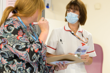 A female clinician in a white coat and face mask is talking to a woman in a patterned dress.
