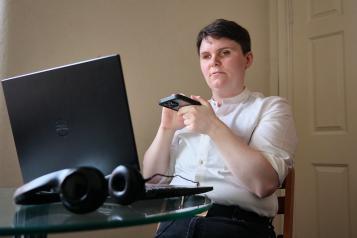 A visually impaired person is sitting at a computer. They are using a screen reader on their mobile phone. They are wearing a white shirt and black trousers.