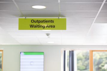 A clinic sign on a green background with black text says 'outpatients waiting area'. There is a window and information board in the background.