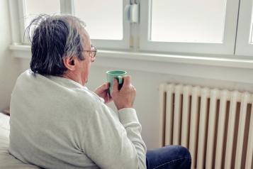 Man in warm sweater with hot tea sitting near radiator trying to warm.
