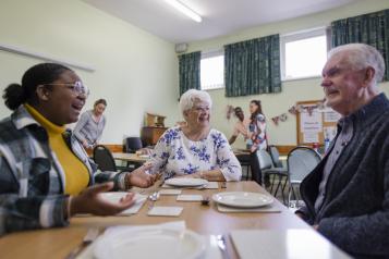 A community church hall running a soup kitchen and warm hub where a diverse group of people in need are having a meal.