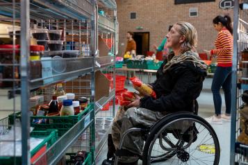 Side view of a volunteer in a wheelchair organising food donations into storage at a food bank in the North East of England. Behind him are his co workers who are working out of focus