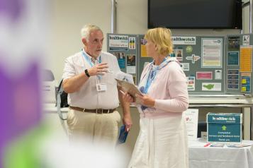A male and female in a hospital corridor. They are in conversation. A Healthwatch information board is behind them.