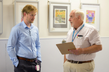 Two men are standing in a clinic having a conversation. The man on the left wears a light blue shirt.  The man on the white wears a blue shirt and Healthwatch lanyard.