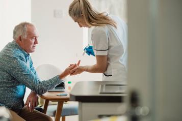 An older man sits in a treatment room with a nurse who is carrying out a blood test on him