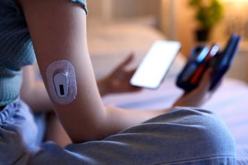 A person is sitting on a bed. They are looking at diabetes monitoring equipment and a mobile phone. They have a monitoring device on their right arm.