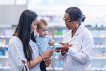 A young mother of Asian descent is picking up a prescription. The woman is holding her baby girl while at the store. She is speaking to a female pharmacist of African descent about the medicine. The medical professional is holding the box of medicine.