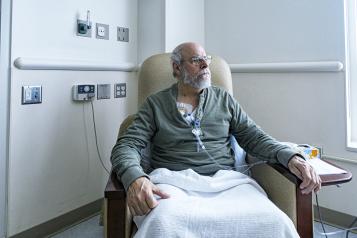A recovering senior adult man colorectal cancer patient is sitting resting comfortably in a hospital cancer ward easy chair while chemotherapy IV drip medicine is administered by an array of medical equipment through a subcutaneous intravenous chemo access port temporarily embedded into his upper chest. 