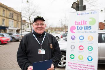 An older man wearing a hat standing in front of a poster promoting Healthwatch