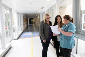 Three people in a hospital corridor. Two people speaking to a nurse