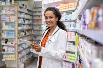 Young female pharmacist. She is wearing a white lab coat and orange shirt. Her hair is tied back. She is holding a tablet.