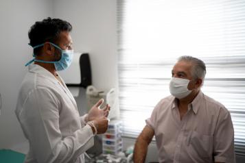 Male patient sitting on a hospital bed and talking to a male doctor. Both are wearing face masks. 