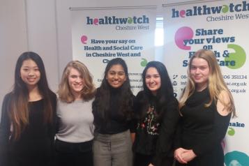 Five teenage girls smiling and standing in front of a Healthwatch banner