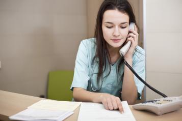 Young female nurse sitting at a desk speaking over the phone while looking at papers