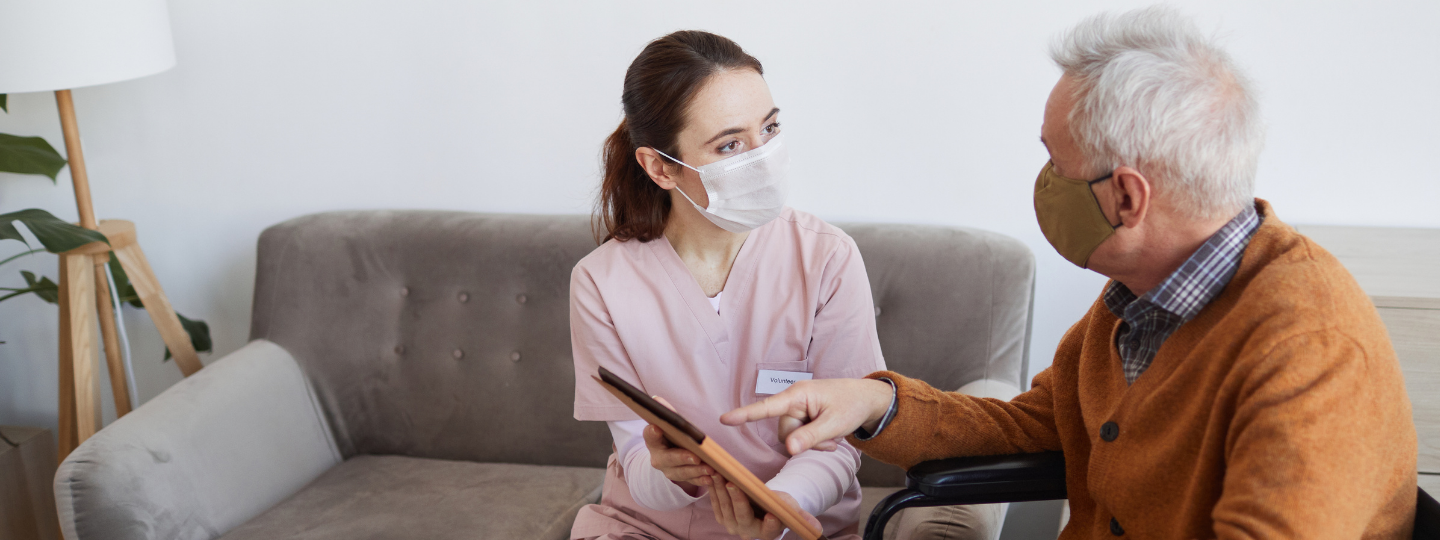 Elderly man using his iPad supported by a young female carer in a mask
