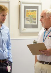 Two men are standing in a clinic having a conversation. The man on the left wears a light blue shirt.  The man on the white wears a blue shirt and Healthwatch lanyard.